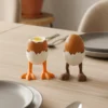 Zwei lustige Eierbecher mit Beinen aus dem 3D-Drucker, dekoriert mit leckeren Eiern auf einem elegant gedeckten, hellen Holztisch.
