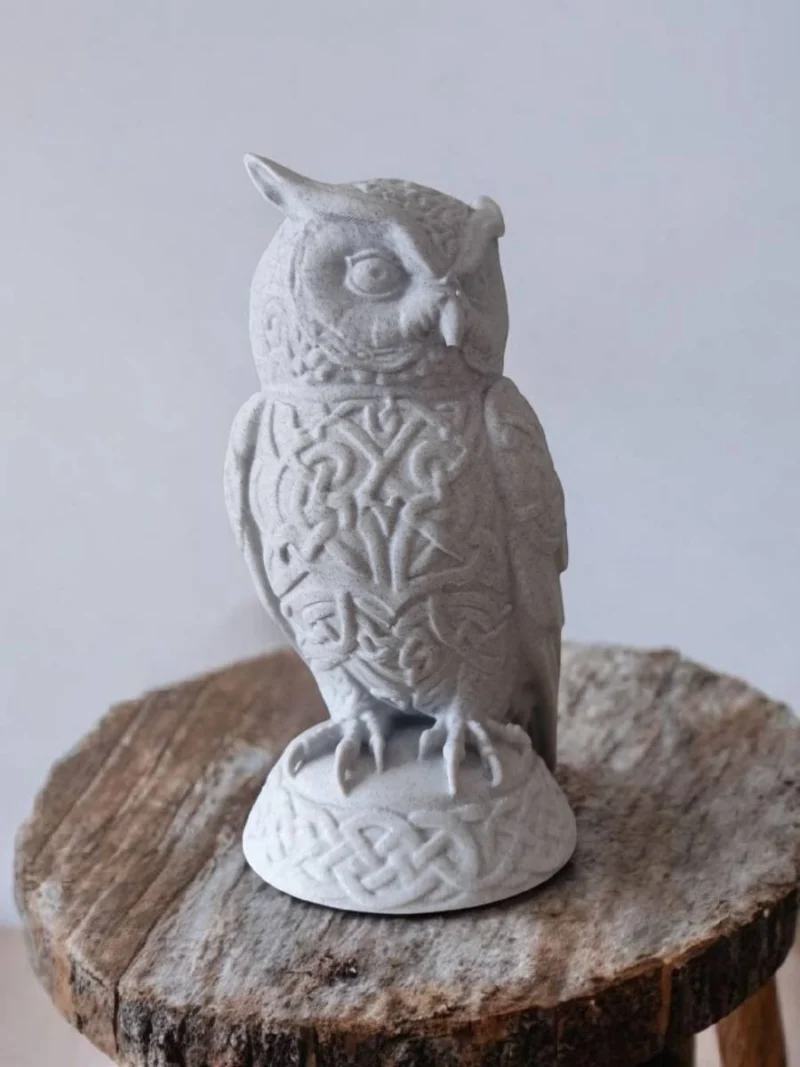 Eine hellgraue Keltische Eulen Skulptur aus dem 3D-Drucker mit detailreichen Ornamenten, die auf einem rustikalen Holztisch dekoriert ist.