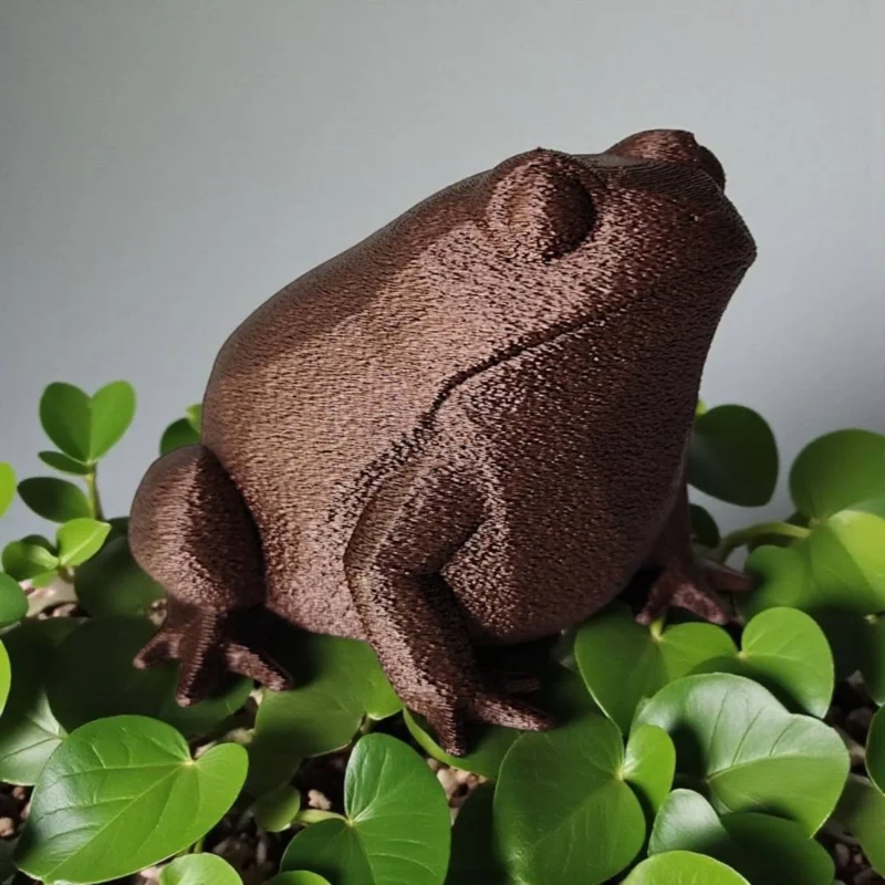 Eine kupferfarbene Low Poly Frosch Skulptur aus dem 3D-Drucker, die stilvoll auf einem Holzregal neben einer kleinen Sukkulente sitzt.