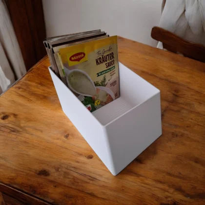 Ein weisser Zutaten Organizer aus dem 3D-Drucker, in dem verschiedene Saucen- und Gewürztüten ordentlich auf einer Holzarbeitsfläche stehen.