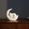 Eine weisse Katze auf Mond Skulptur aus dem 3D-Drucker, die friedlich schlafend auf einem Regal aus dunklem Holz dekoriert ist.
