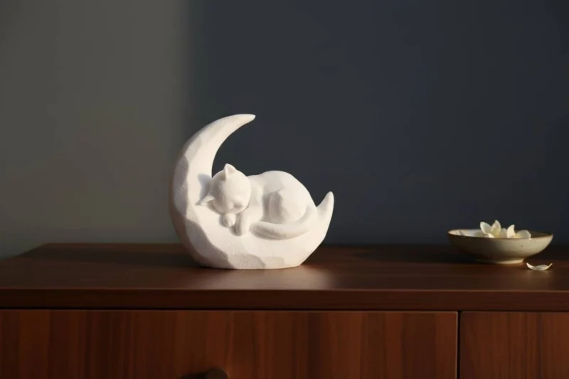 Eine weisse Katze auf Mond Skulptur aus dem 3D-Drucker, die friedlich schlafend auf einem Regal aus dunklem Holz dekoriert ist.