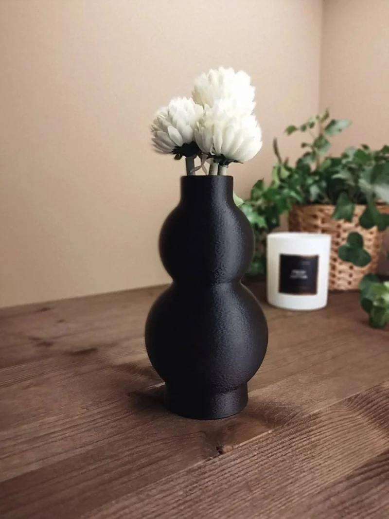 ine schwarze geschwungene Design Vase aus dem 3D-Drucker mit edler Fuzzy-Skin-Struktur, dekoriert mit weissen Blumen auf einem dunklen Holztisch.