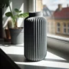 Eine schwarze minimalistische gerippte Vase aus dem 3D-Drucker, die stilvoll in einem hellen, sonnigen Raum auf einem glatten Holzboden präsentiert wird.