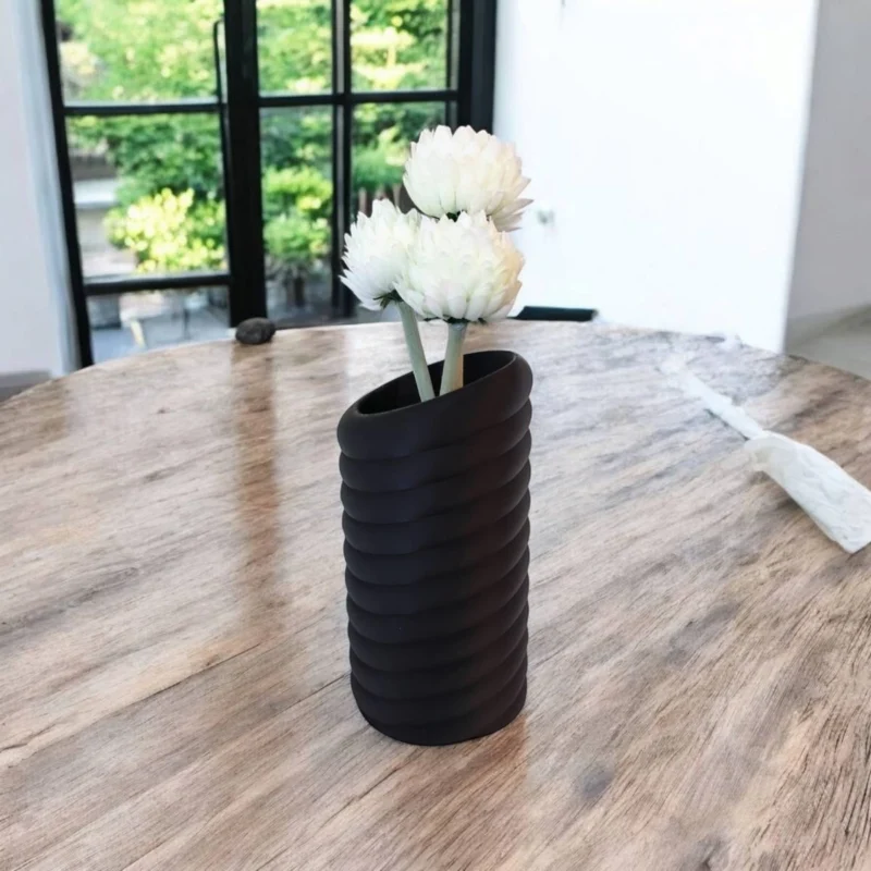 Eine schwarze moderne Swirl Vase aus dem 3D-Drucker, die stilvoll mit weissen Blumen auf einem rustikalen Holztisch dekoriert ist.