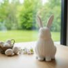 Eine weisse Osterhase Deko Figur aus dem 3D-Drucker, die stilvoll auf einem hellen Holztisch neben pastellfarbenen Ostereiern steht.