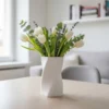 Eine weisse moderne gedrehte Vase aus dem 3D-Drucker (wie in grafik_13.png abgebildet), die stilvoll mit frischen Frühlingsblumen auf einem Holztisch dekoriert ist.