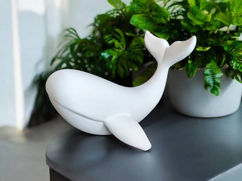 Detailaufnahme der sanften, fliessenden Linien und der hochwertigen, matten Oberfläche der minimalistischen Wal Skulptur vor grünen Pflanzen.